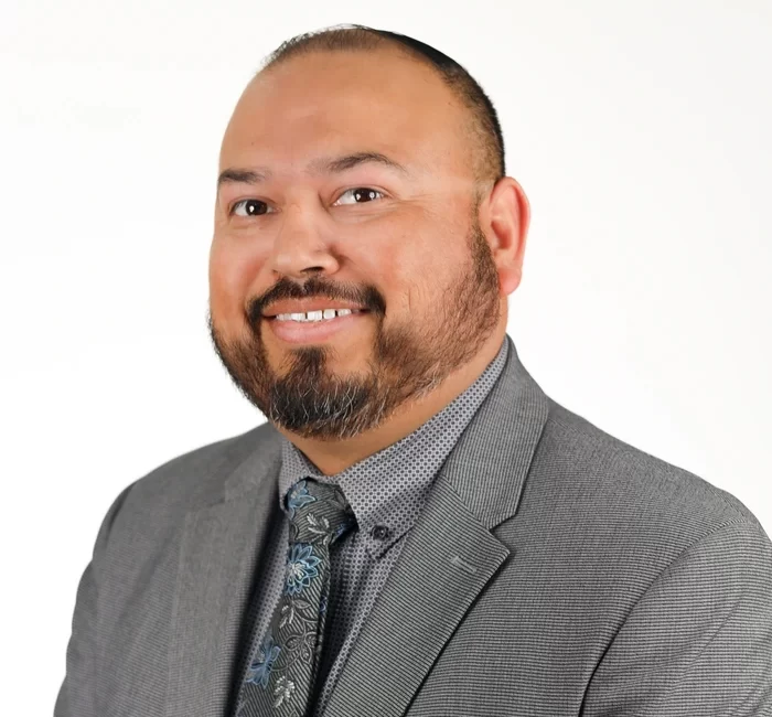 headshot with white background of hispanic man with dark goatee wearing a grey suit