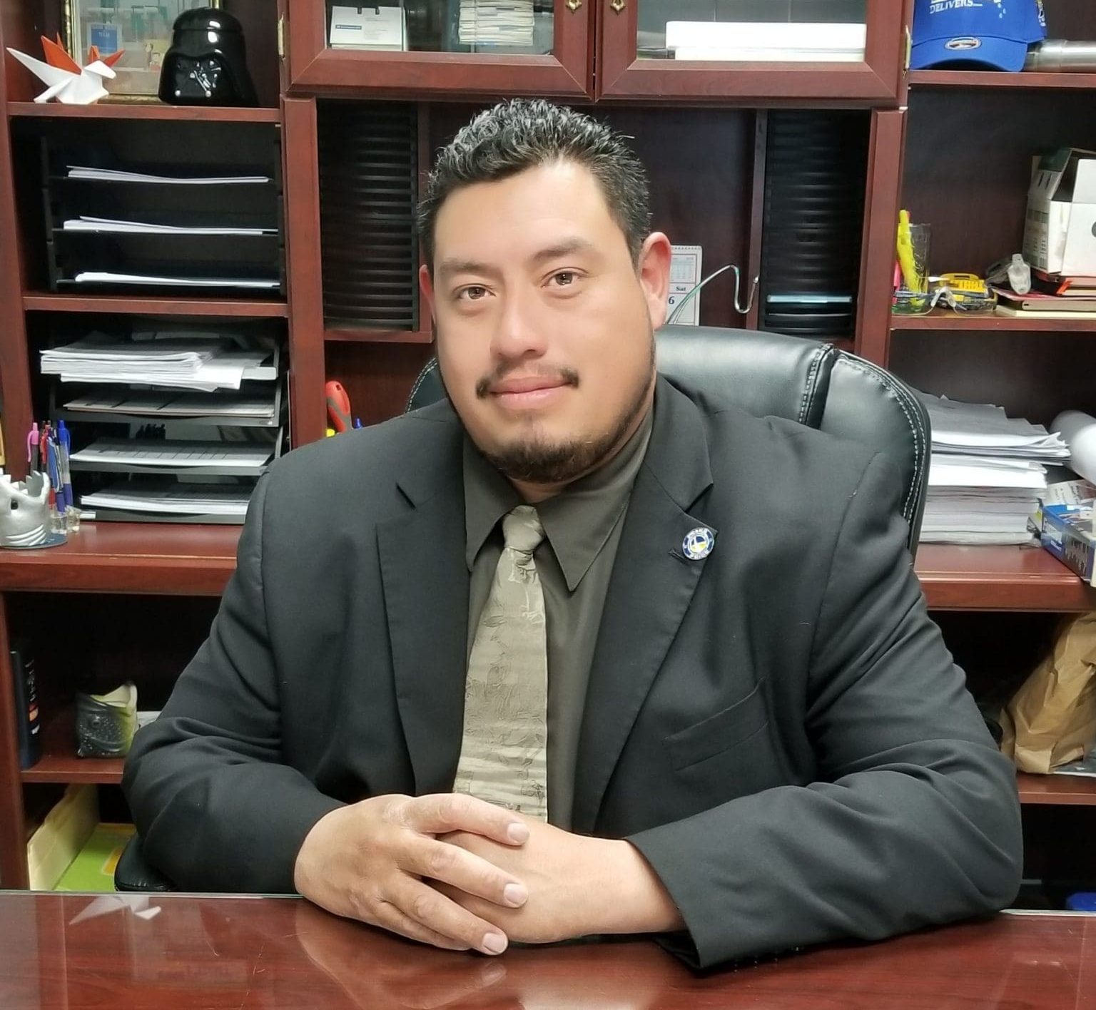 hispanic man wearing dark suit seated at dark wooden desk with dark wooden bookcase behind him holding various papers and office equipment